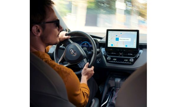 A man in an orange jumper turns rights as he drives a Toyota car. The car's multimedia system displays a software update.